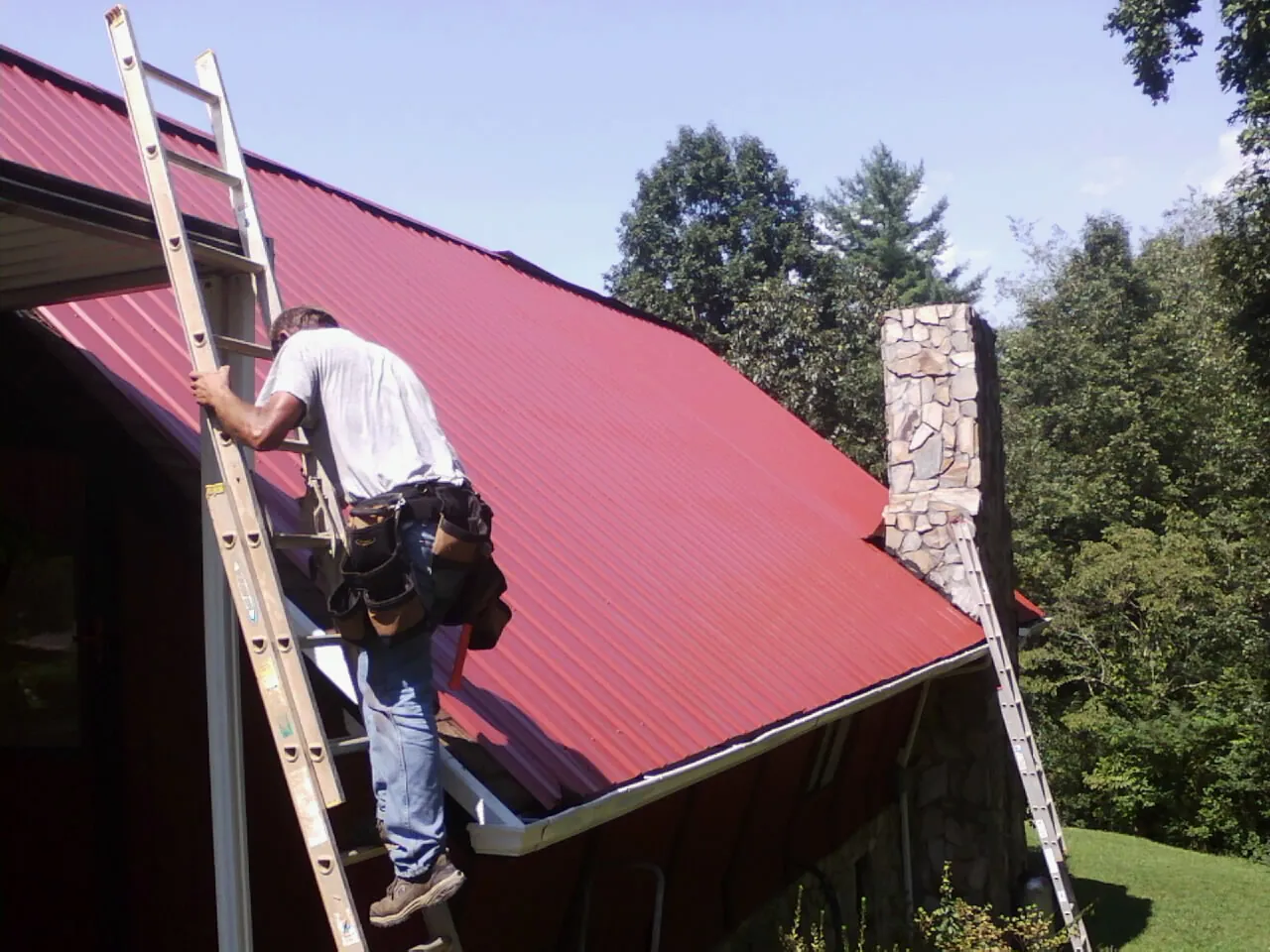 Roofer on a ladder beside a red metal roof and stone chimney — Soffit & Fascia Repair by Roofing Brothers in Newton Highlands