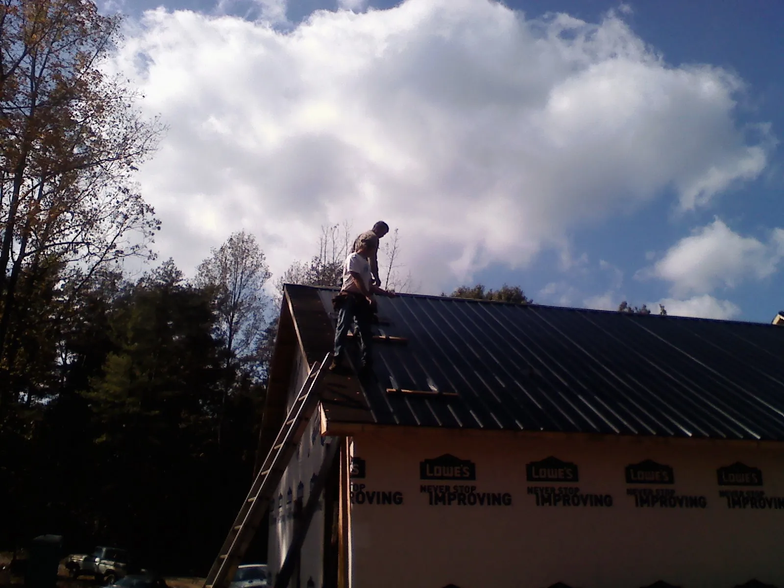 Roofer fastening dark metal panels high against a cloudy sky, from a Roof Inspection job in Newton Highlands